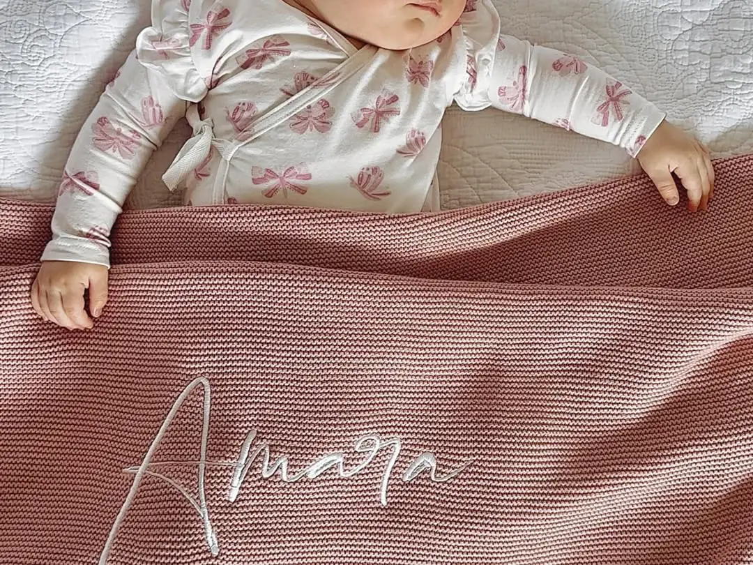 A baby resting under a dusty-pink knitted blanket embroidered with the name Amara