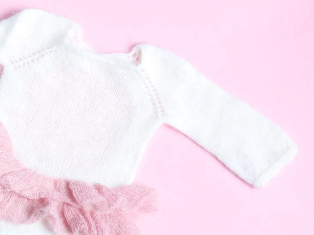 A white knitted baby onesie with pink ruffles laid out on a soft pink background
